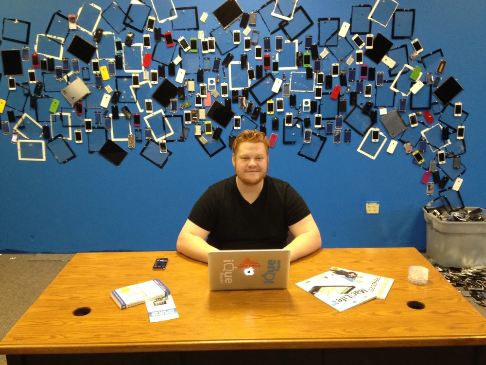 Young Morgan McKell at a desk in front of a wall of repaired phone screens at iQue Repair, the cell phone repair company he co-founded in 2011